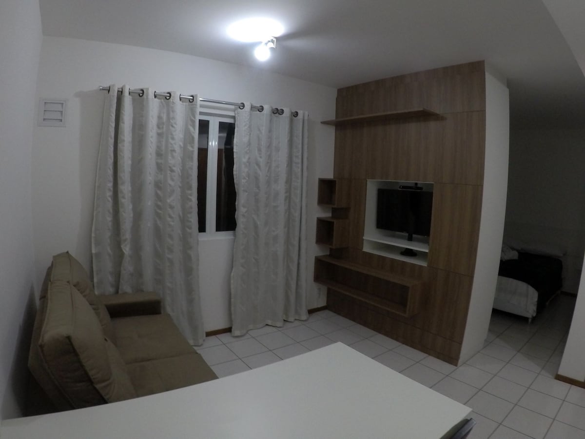 A compact living area is featured with light-colored tiled flooring. A soft couch is positioned next to a wall unit that includes a mounted television. Sheer curtains frame a window, allowing natural light to brighten the space.
