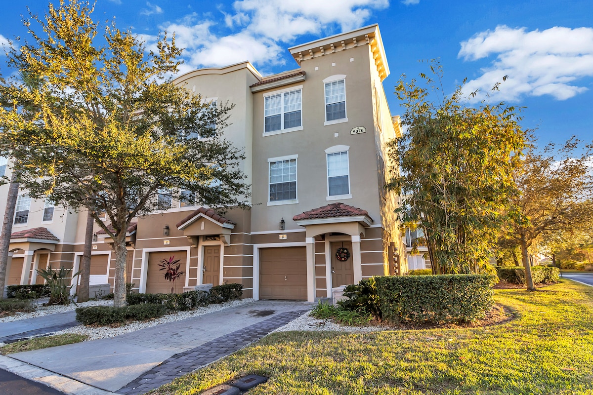 The exterior of the three-story townhome features a well-maintained facade with multiple windows under a blue sky. The entrance displays a charming wreath on the door, framed by landscaped shrubs and trees. A paved walkway leads to the front entrance, enhancing the welcoming appearance.
