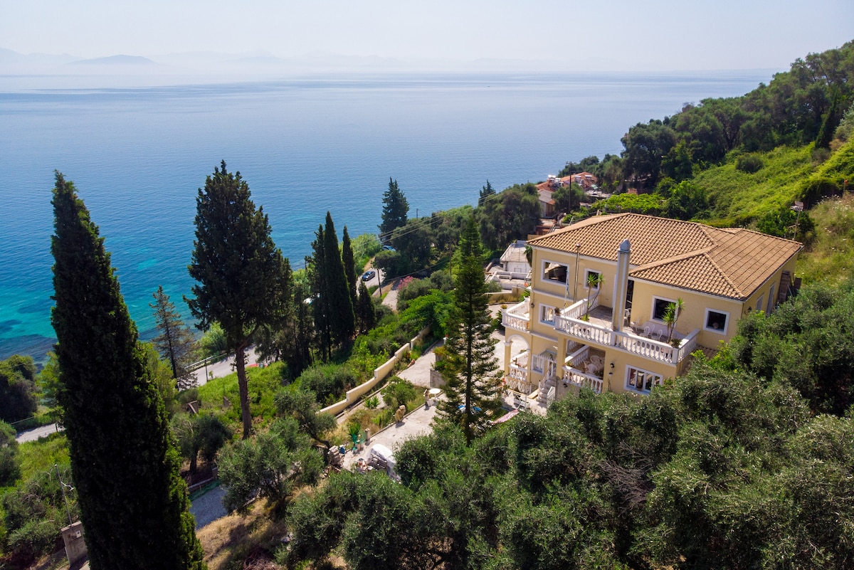 A spacious apartment is positioned on a hillside, featuring a terracotta roof. Surrounding trees and shrubs contribute to a natural ambiance. The Ionian Sea extends in the background, offering wide views of the blue waters and distant landforms, creating a calm and serene setting.