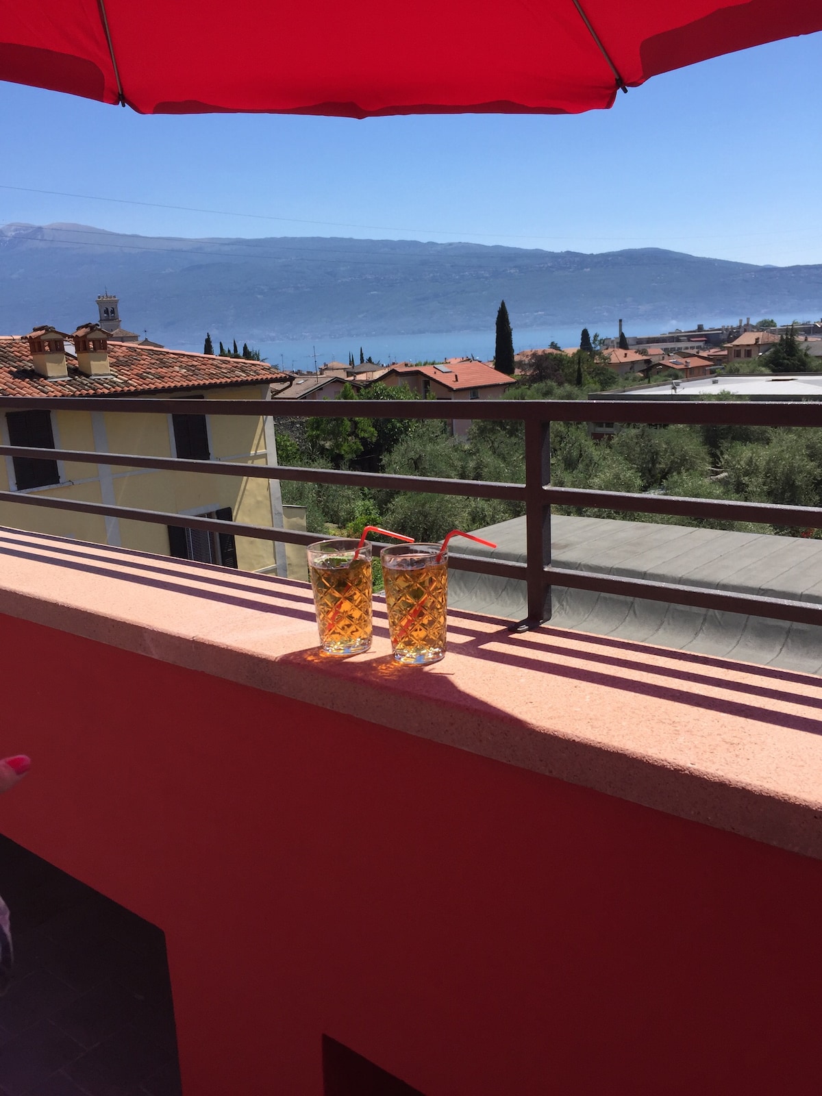Two glasses with colorful straws are positioned on a ledge, overlooking a panoramic view of the landscape. Rolling hills and a lake are visible in the distance, framed by greenery and rooftops. A vibrant red umbrella provides shade.