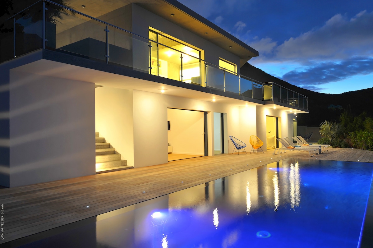 The contemporary villa exterior is illuminated at night, showcasing large glass doors that lead inside. A heated pool reflects soft lights, while wooden decking surrounds the water. Lounge chairs are arranged nearby, enhancing the inviting outdoor space.