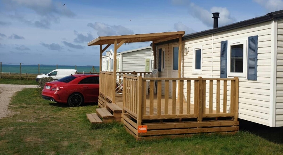 A wooden terrace is connected to a mobile home, featuring a railing for added safety. The structure faces the sea, with a car parked nearby on the gravel driveway. Grass surrounds the area, with a clear view of the ocean in the background.