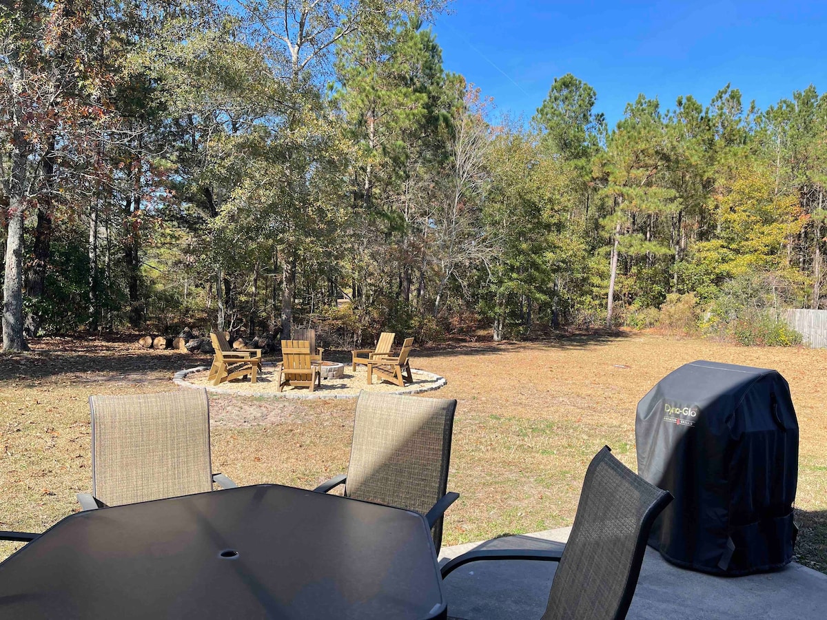 A spacious backyard is visible, featuring a circular firepit surrounded by wooden chairs. A patio table is set nearby, and a grill is positioned on the patio surface. Lush trees provide a natural backdrop under a clear blue sky.