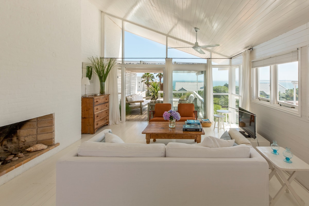 A bright and airy living space is adorned with white walls and a high ceiling, enhancing the feeling of openness. Large windows invite natural light and provide views of the ocean. A comfortable sofa sits opposite a wooden coffee table, creating a casual seating area.