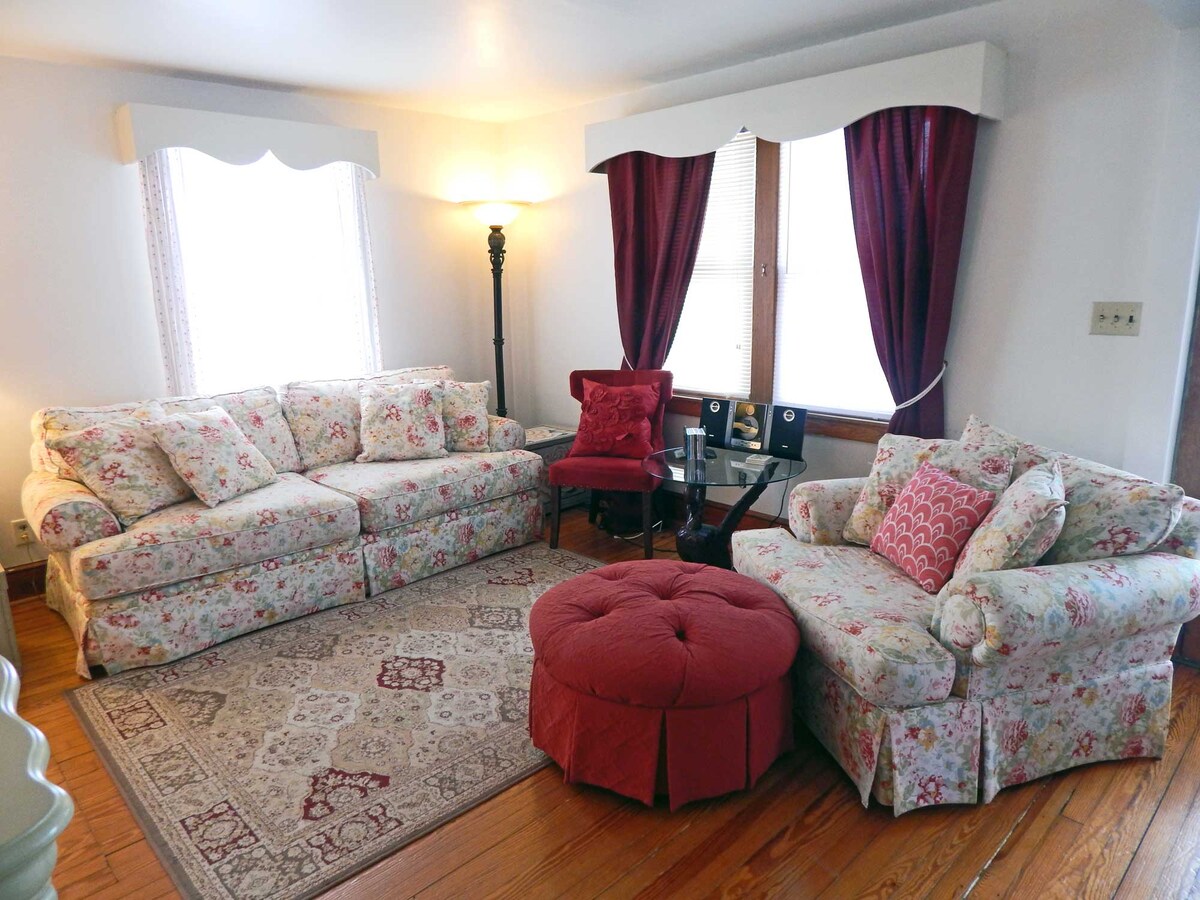 A living room area is presented with floral-patterned sofas and armchairs, complemented by a round ottoman. Natural light filters through the windows, adorned with rich, burgundy drapes. A small table sits between the seating, enhancing the cozy ambiance of the space.