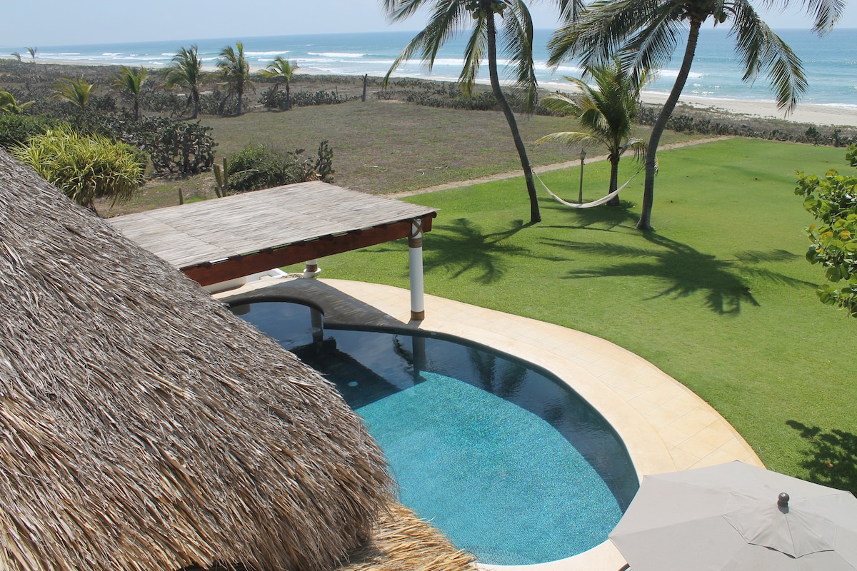 A serene view of the oceanfront villa is captured, showcasing a curved pool surrounded by lush green grass. Coconut palms sway gently in the breeze, and a hammock can be seen positioned near the water's edge, enhancing the tranquil atmosphere.