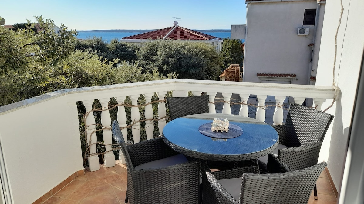 A balcony features a round table surrounded by four wicker chairs, offering a serene view of the sea. Greenery surrounds the area, with a backdrop of rooftops and distant water, creating a peaceful outdoor setting.