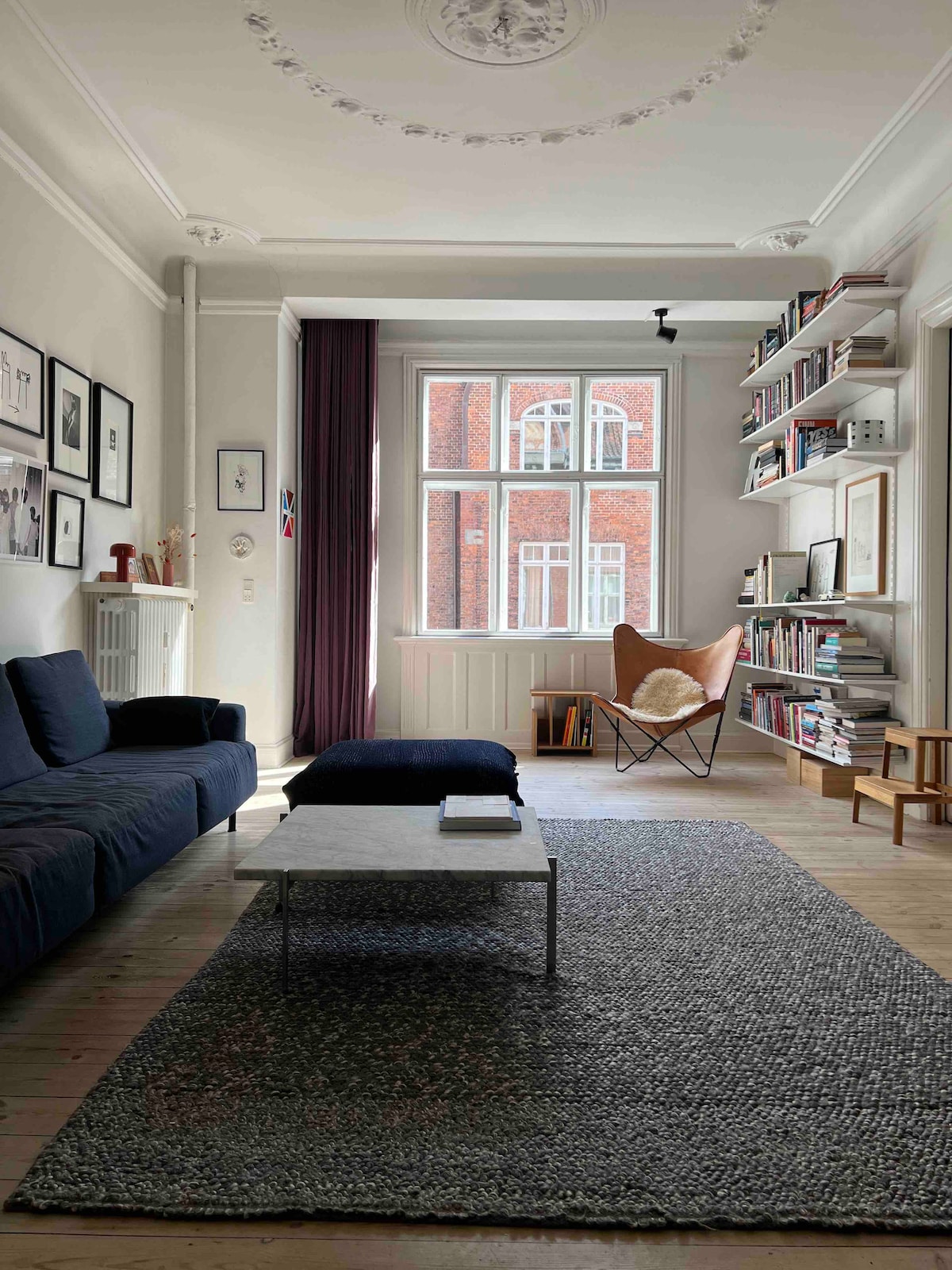 The living area is filled with natural light from large windows. A dark blue sofa is positioned near a textured rug, while a low coffee table rests in front. A cozy reading nook features a butterfly chair and shelves filled with books, enhancing the inviting ambiance.