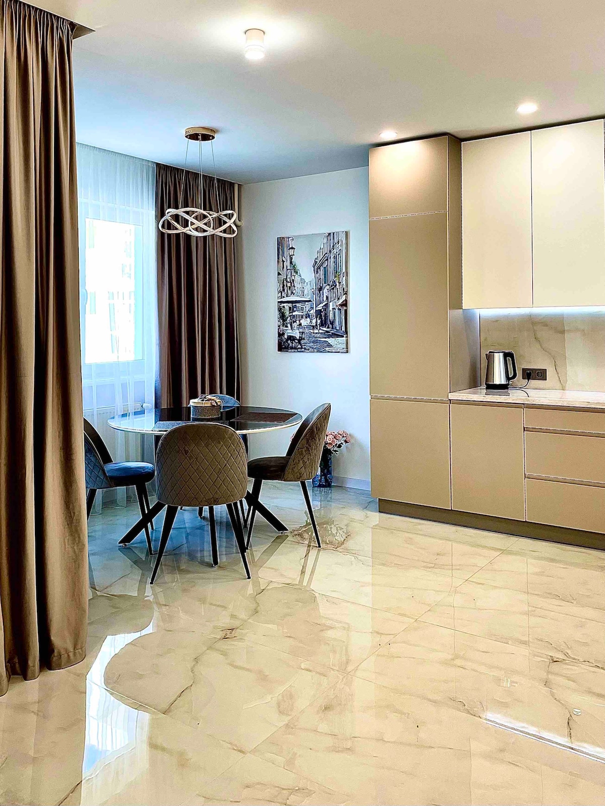 A modern kitchen area features a round dining table surrounded by four upholstered chairs. Soft lighting illuminates the space, highlighting the glossy tiled floor and neutral-colored cabinetry. A framed artwork is displayed on the wall, adding a touch of character to the room.