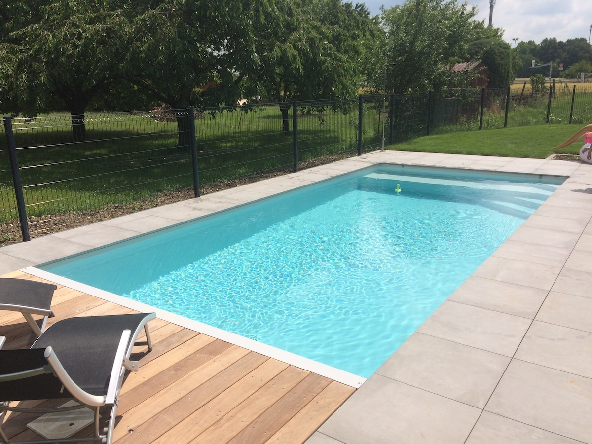A rectangular swimming pool is surrounded by a sleek concrete border. Wooden decking complements the pool area, providing space for lounge chairs. Lush green grass and trees are visible in the background, contributing to a serene outdoor environment.