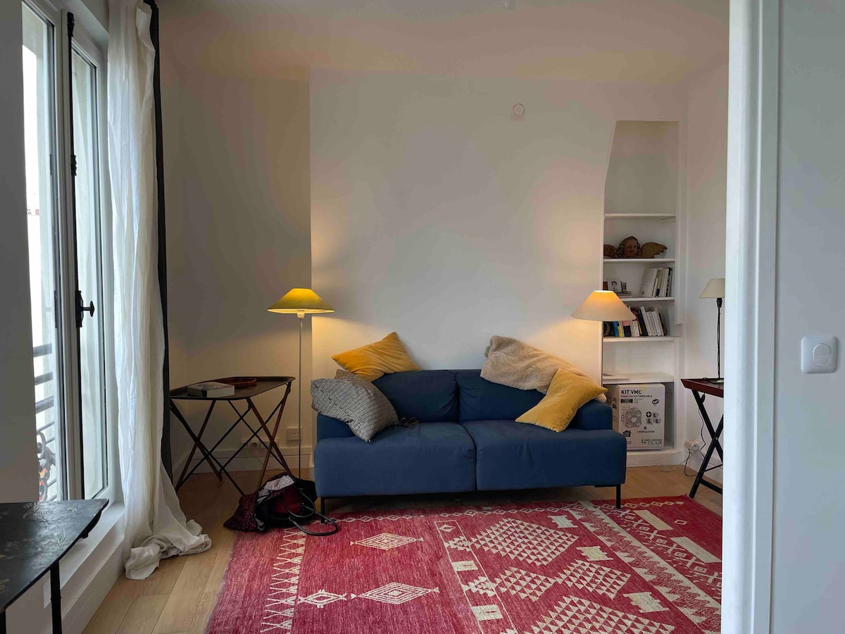 The cozy living area features a blue sofa adorned with two yellow cushions, complemented by two side tables and a bookshelf. Soft lighting is provided by two lamps. A vibrant red rug anchors the space, while natural light filters in through large windows.