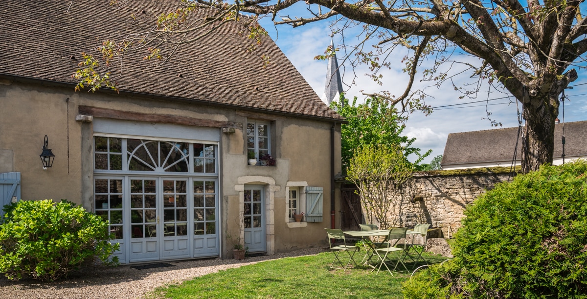 A charming countryside home is presented with a spacious garden area. French doors enhance the entrance, while a tree provides shade over a small outdoor seating arrangement with metal furniture. Lush greenery complements the scenic view, contributing to a tranquil environment.