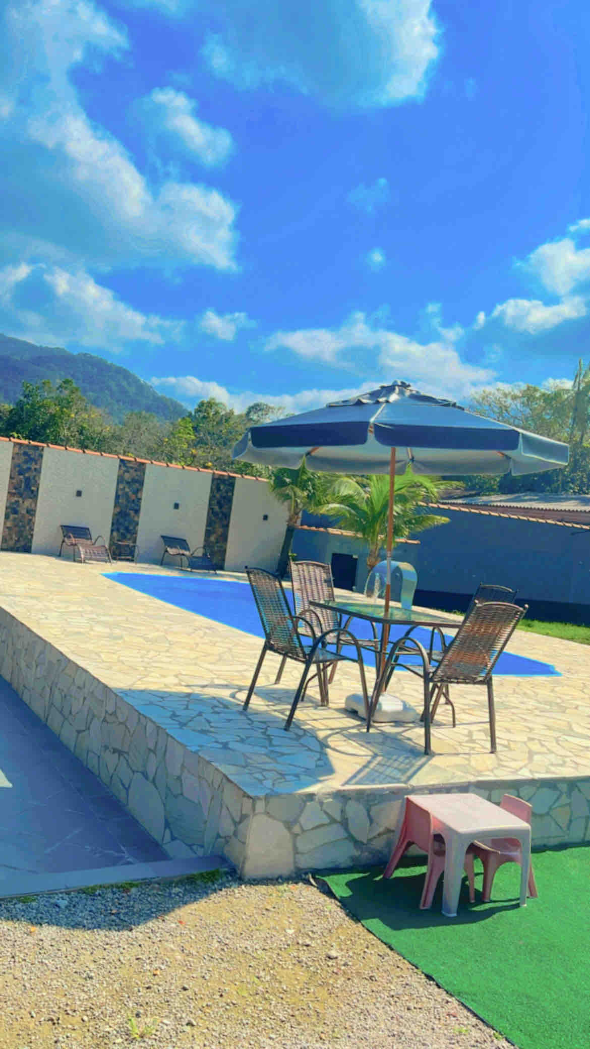 A well-maintained swimming pool is surrounded by a stone patio, featuring a shaded seating area with a table and chairs. A large umbrella offers protection from the sun, while greenery is visible in the background, framed by mountain views and a clear blue sky.