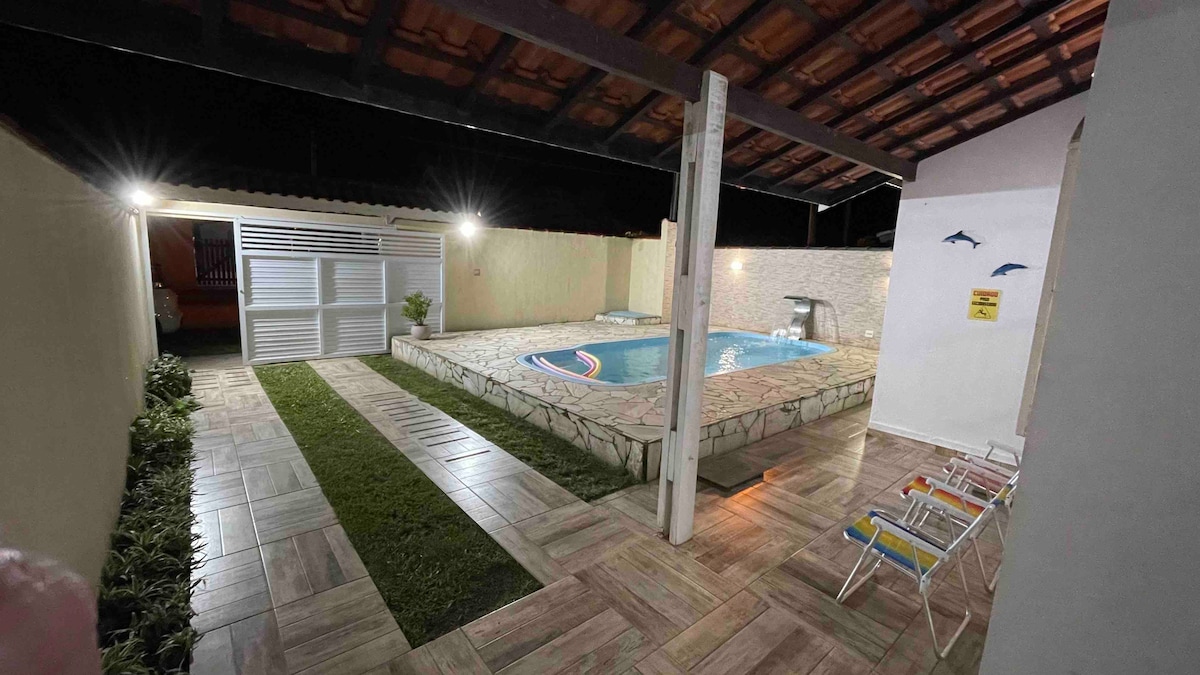 The outdoor area features a well-maintained swimming pool surrounded by a tiled deck. Three beach chairs and green grass are positioned nearby, and soft lighting illuminates the space, creating a welcoming atmosphere for evening gatherings.
