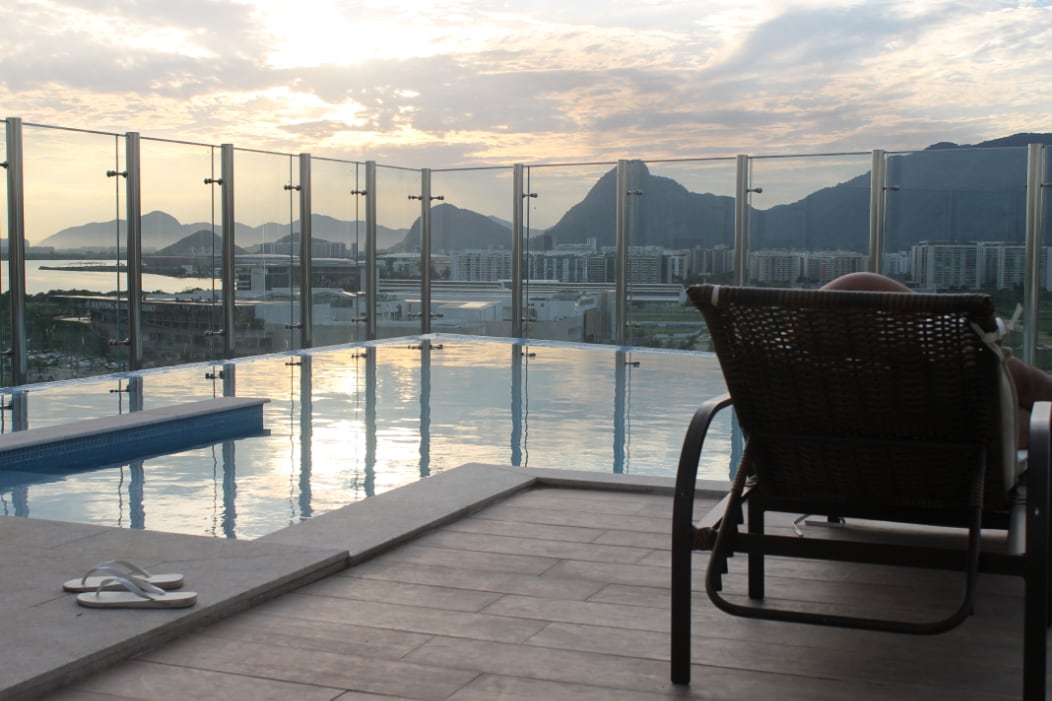 A serene rooftop area is depicted, featuring an infinity pool that reflects the surrounding landscape. Comfortable lounge chairs are positioned near the pool, with mountains in the background and a subtle sunset illuminating the sky.