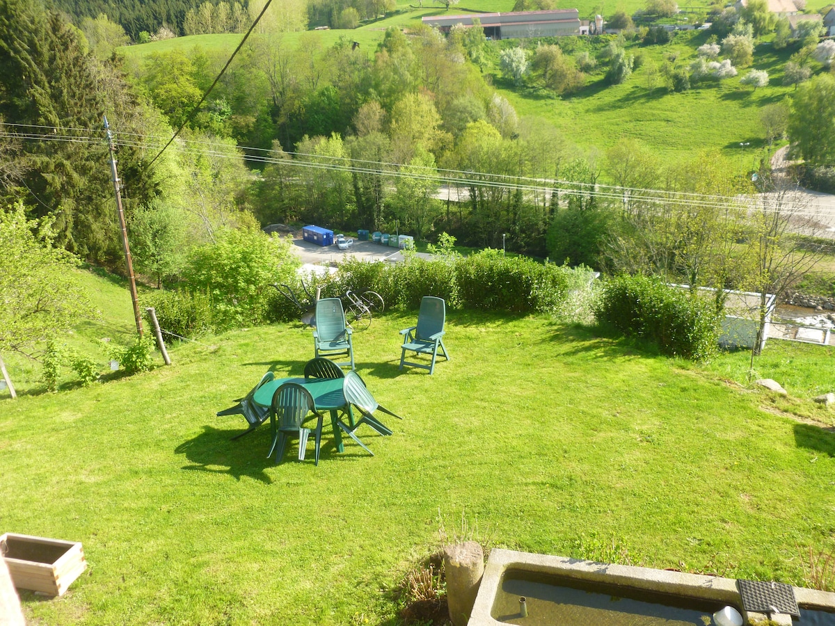 A green outdoor space is depicted, featuring a circular arrangement of plastic chairs surrounding a table. Lush grass covers the area, bordered by trees and shrubs. In the background, a winding road and distant hills can be seen, along with a few scattered bicycles.