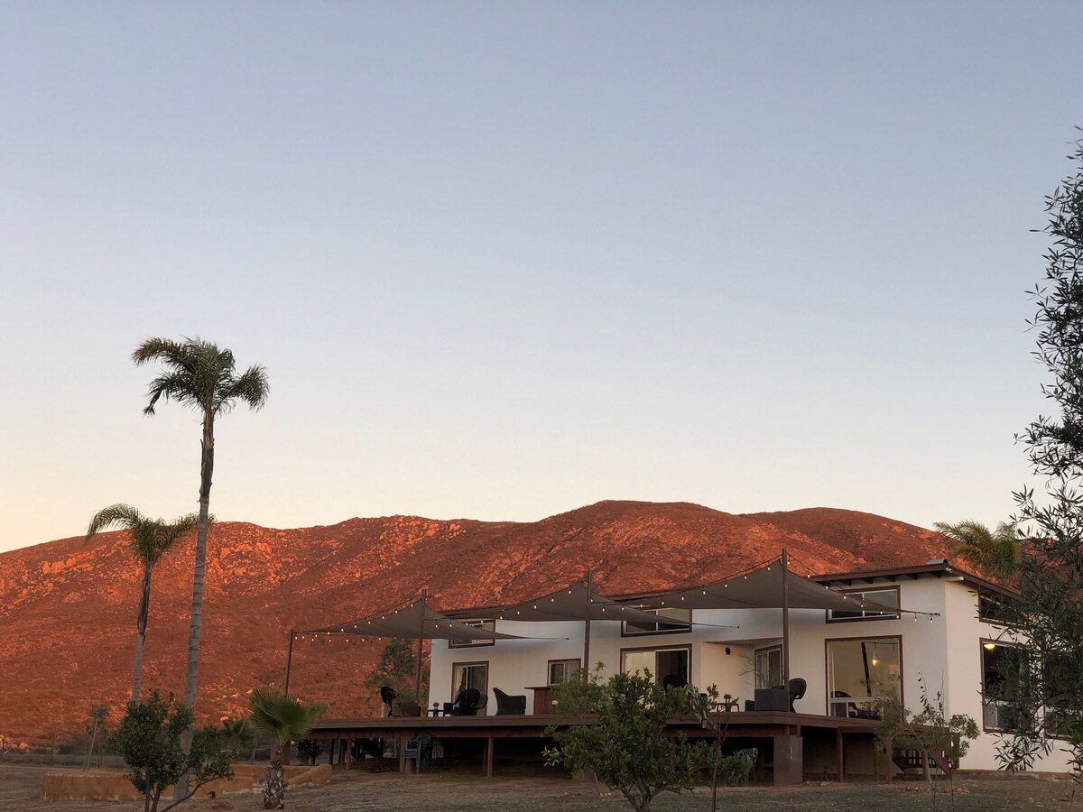 The exterior of the house features a spacious deck shaded by large awnings, providing seating areas for relaxation. Palm trees frame the setting, creating a serene atmosphere against the backdrop of a gently rising mountain, bathed in warm evening light.