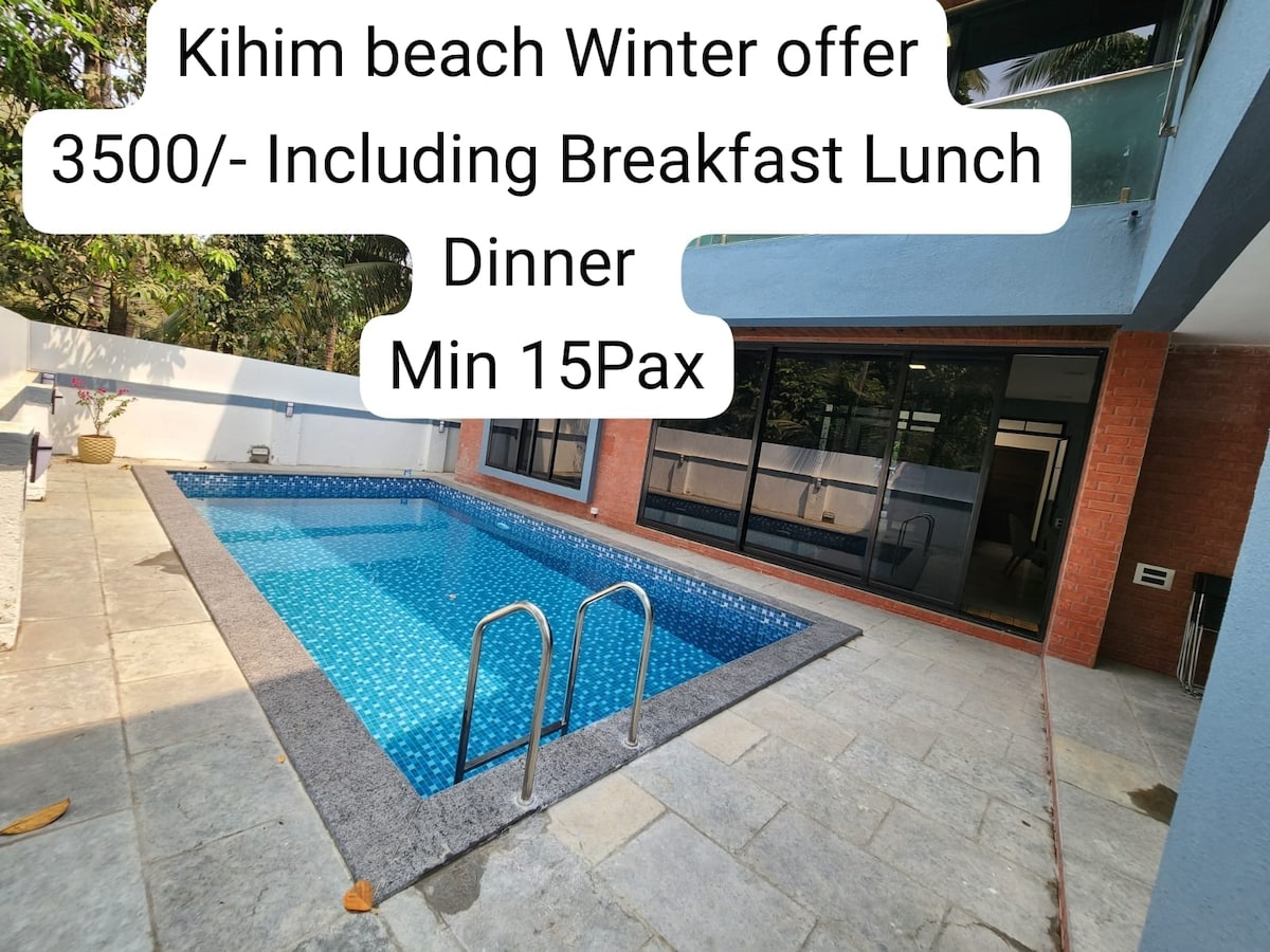 A clear blue swimming pool is surrounded by a stone patio, dotted with a few fallen leaves. A shaded area is visible in the background, offering a view of greenery and a modern building facade. The inviting water reflects sunlight, creating a tranquil ambiance.