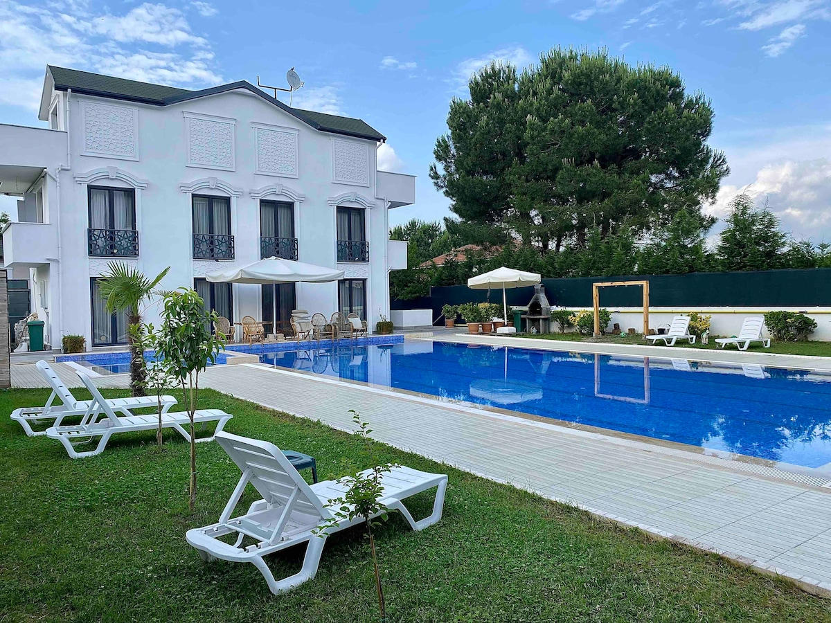 A spacious outdoor area features a large swimming pool surrounded by lounge chairs and greenery. Flanking the pool, a modern two-story villa displays multiple windows with decorative balconies, while a shaded seating area is positioned nearby for relaxation.