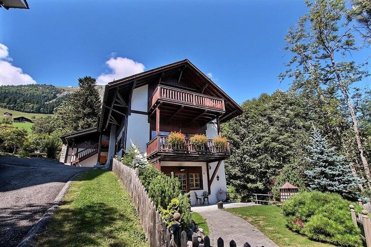A charming chalet is set against a backdrop of green hills and blue skies. The building features a wooden exterior with balconies adorned with vibrant flowers. A pathway lined with bushes leads to the entrance, enhancing the inviting atmosphere of the property.