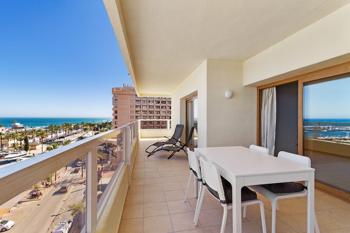 A private terrace is shown with a dining table and four chairs. A pair of lounge chairs is positioned nearby, overlooking the marina and sea. Large glass doors provide access to the interior, with a clear view of the bright sky and waterfront.