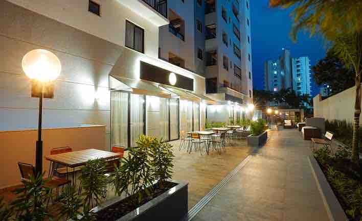 An outdoor area is illuminated by soft lighting, featuring multiple patio tables and chairs arranged along a paved walkway. Lush greenery adds a touch of nature, while a modern building stands in the background, reflecting the evening sky.