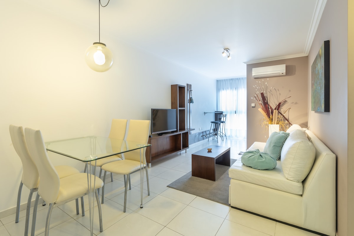 A bright living area features a glass dining table with four upholstered chairs. A white sofa is arranged beside a low coffee table on a textured rug. Natural light flows in through large windows, highlighting the minimalistic decor and a wooden entertainment unit.