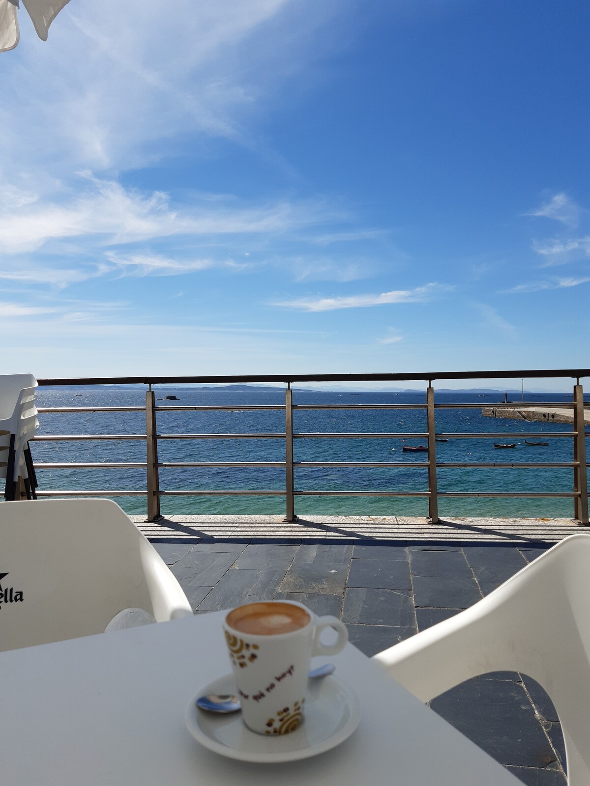 A table is set on a terrace overlooking the ocean. A small cup of coffee is placed on the table, next to a silver saucer. The sea shimmers beneath a clear blue sky, with distant boats visible on the horizon.