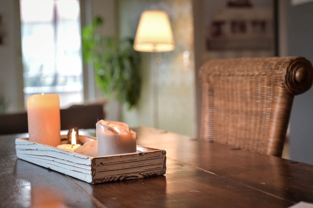 A tray with three assorted candles sits on a wooden dining table, with a cozy lamp in the background illuminating the space. A woven chair is positioned nearby, complemented by a soft green plant visible through a window.