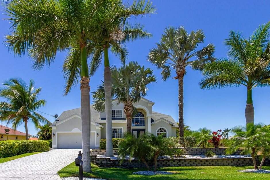 The exterior of the villa features a grand entrance with a double-height window, flanked by palm trees and vibrant landscaping. A paved driveway leads up to the house, enhancing the tropical ambiance. Bright blue skies create a striking contrast against the light-colored facade.
