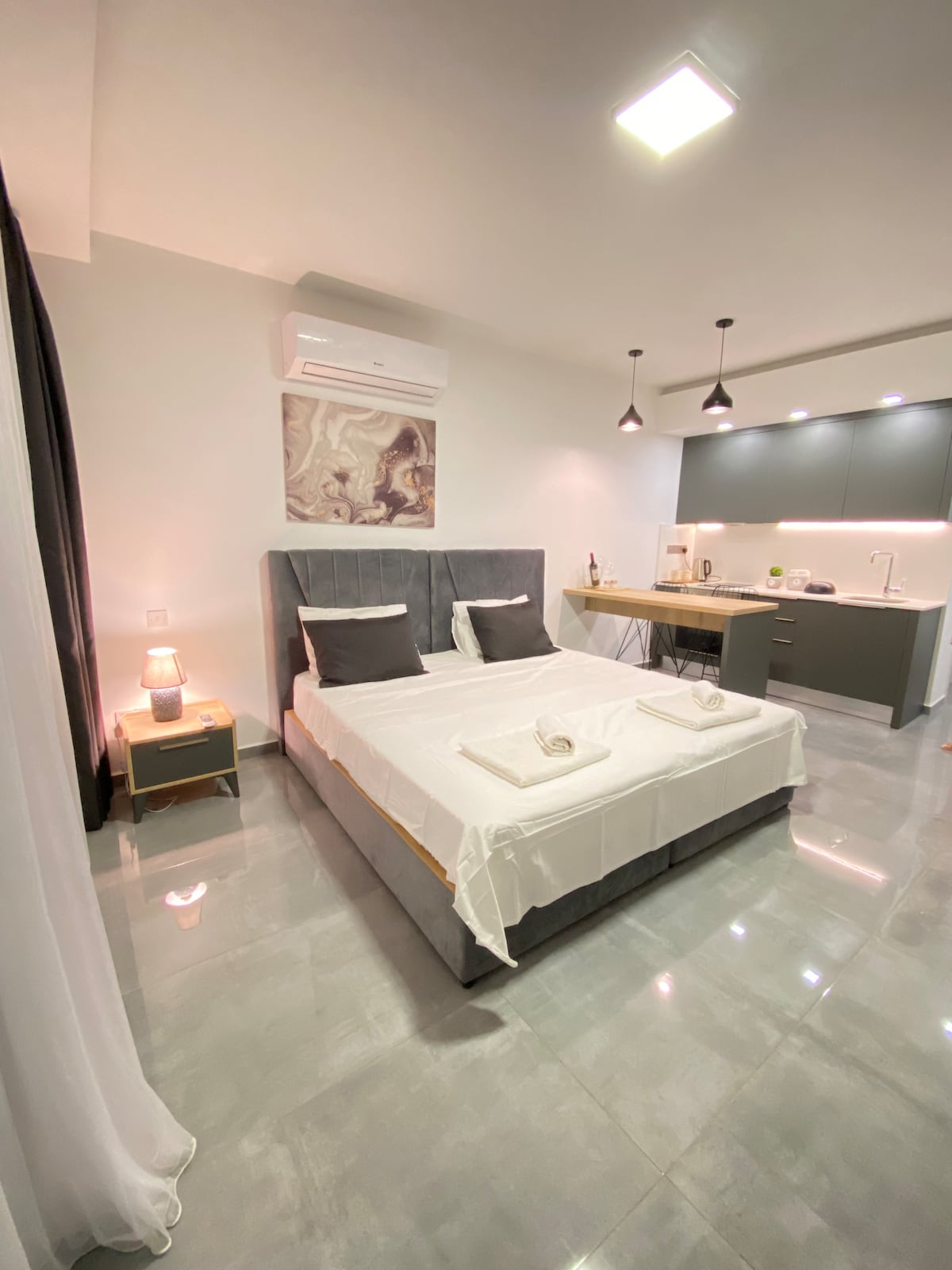 A modern studio bedroom features a spacious bed dressed in neutral tones, complemented by two bedside lamps. A kitchenette with sleek cabinetry is visible in the background, while soft lighting enhances the overall ambiance. Light-colored tiles cover the floor, creating a clean and open feel.