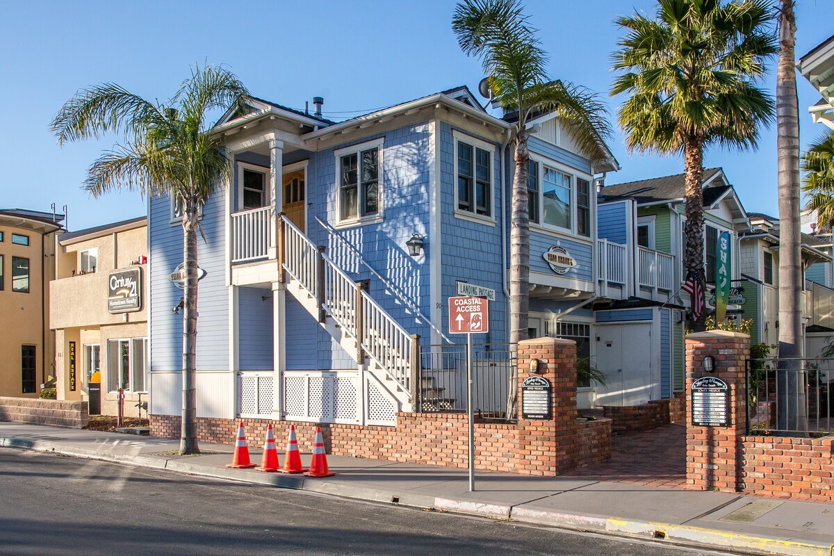 A charming two-story blue building features a welcoming entrance accessed by a set of stairs. Lush palm trees frame the facade, and brick accents provide a sturdy base. Nearby signage indicates directions, while vibrant colors and clear skies enhance the inviting appearance of the property.