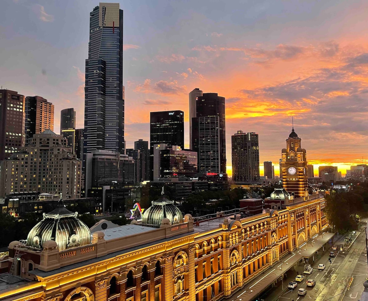 The image captures a vibrant sunset over Melbourne, featuring the historic Flinders Street Station with its illuminated façade in the foreground. Modern skyscrapers rise in the background, and the sky is painted with warm hues of orange and purple, creating a captivating city skyline.