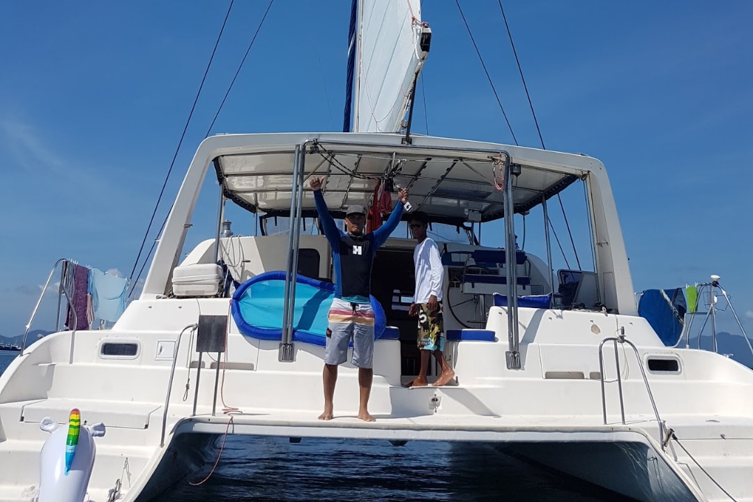 The rear view of a modern catamaran shows spacious outdoor seating and a sturdy frame. The sailing area is equipped with clear windows, while colorful beach towels are draped over the sides. The boat is anchored in tranquil waters under a clear blue sky.