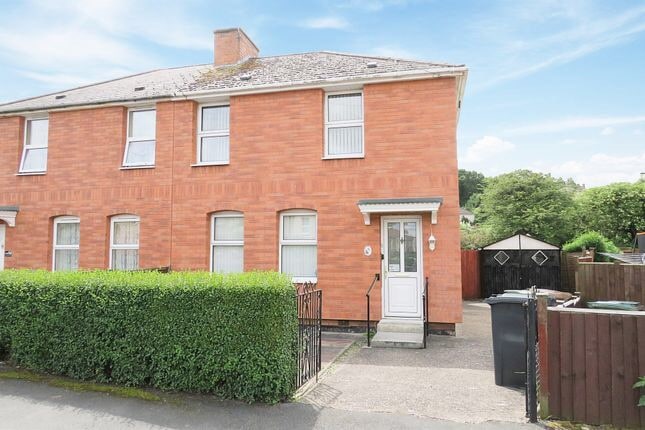 A two-story brick house is shown, featuring large windows that allow ample light. A small front garden with trimmed bushes frames the entrance. The pathway leads directly to a white front door, providing a welcoming entry point to the home.