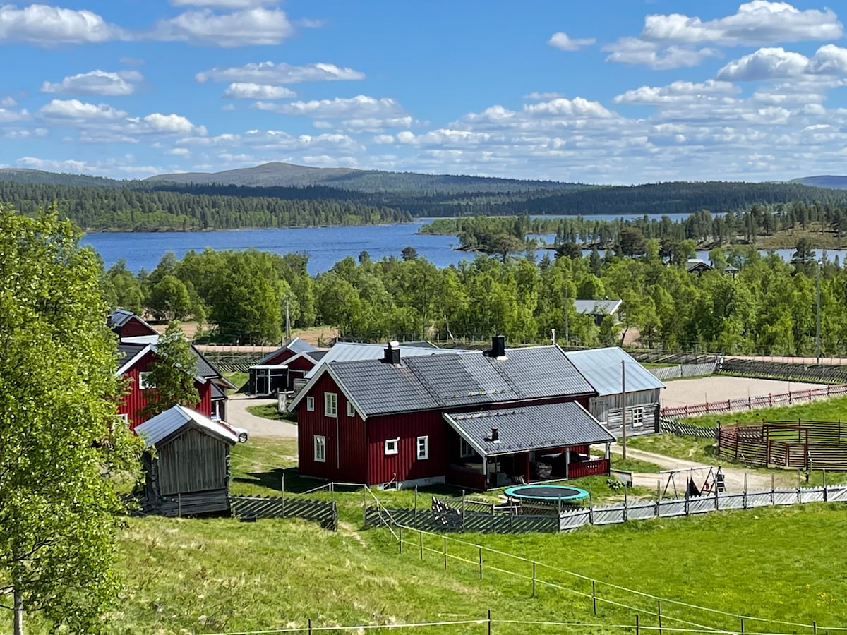A scenic view captures several traditional red wooden buildings set against a backdrop of lush greenery and a tranquil lake. In the foreground, a large grassy area features a trampoline. The sky above is bright with scattered clouds, enhancing the natural beauty of the location.