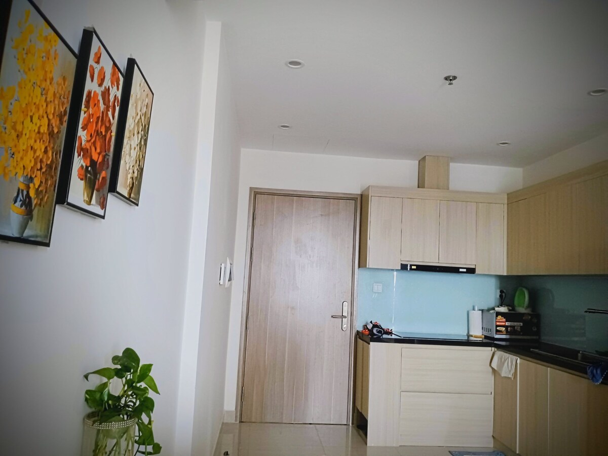 A well-lit kitchen area features neutral cabinetry paired with a light blue backsplash. A solid wooden door leads to another space, while two floral paintings adorn the wall. A small green plant adds a touch of nature to the scene.