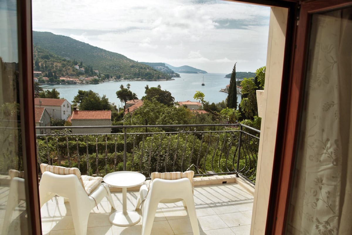 A terrace features a small round table with two white chairs, offering a clear view of the sea and surrounding landscape. The scene includes distant hills and homes, with a sailboat visible in the water. Large glass doors allow natural light to enter the space.