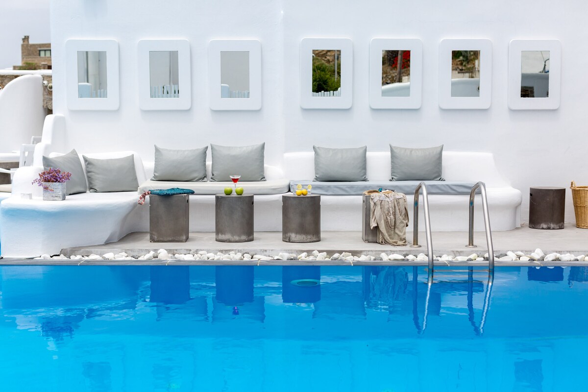 A neatly arranged lounging area beside a swimming pool features a long white bench adorned with gray cushions. Stone tables are positioned throughout, holding decorative items and refreshments. The vibrant blue water of the pool reflects the sunlight, enhancing a serene ambiance.