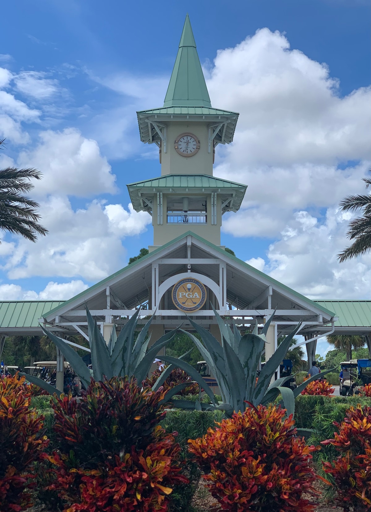 A tall clock tower with a spire is prominent, surrounded by vibrant tropical plants. The structure features a large circular sign indicating the PGA, while clear skies and fluffy clouds form the backdrop.