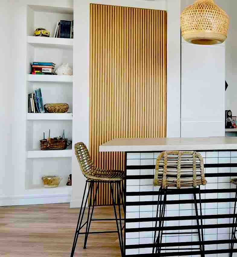 A modern kitchen area features a stylish bar with natural wood and black metal accents. Two high stools with woven seats are positioned at the bar. Behind, a natural wood panel adds texture, while shelves display neatly arranged books and decorative items.