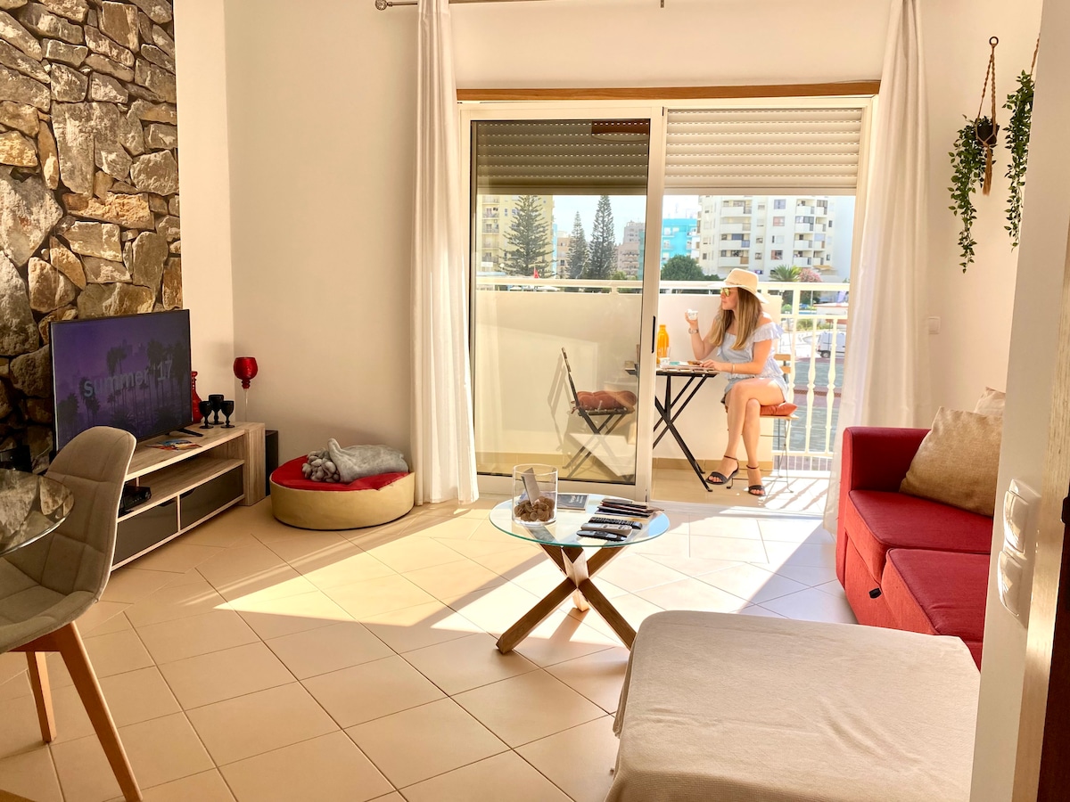 A sunny living area features a red sofa and a glass coffee table. A flat-screen TV is positioned on a media unit. A small balcony can be seen through the sliding glass doors, with a table and two chairs set up for outdoor relaxation.