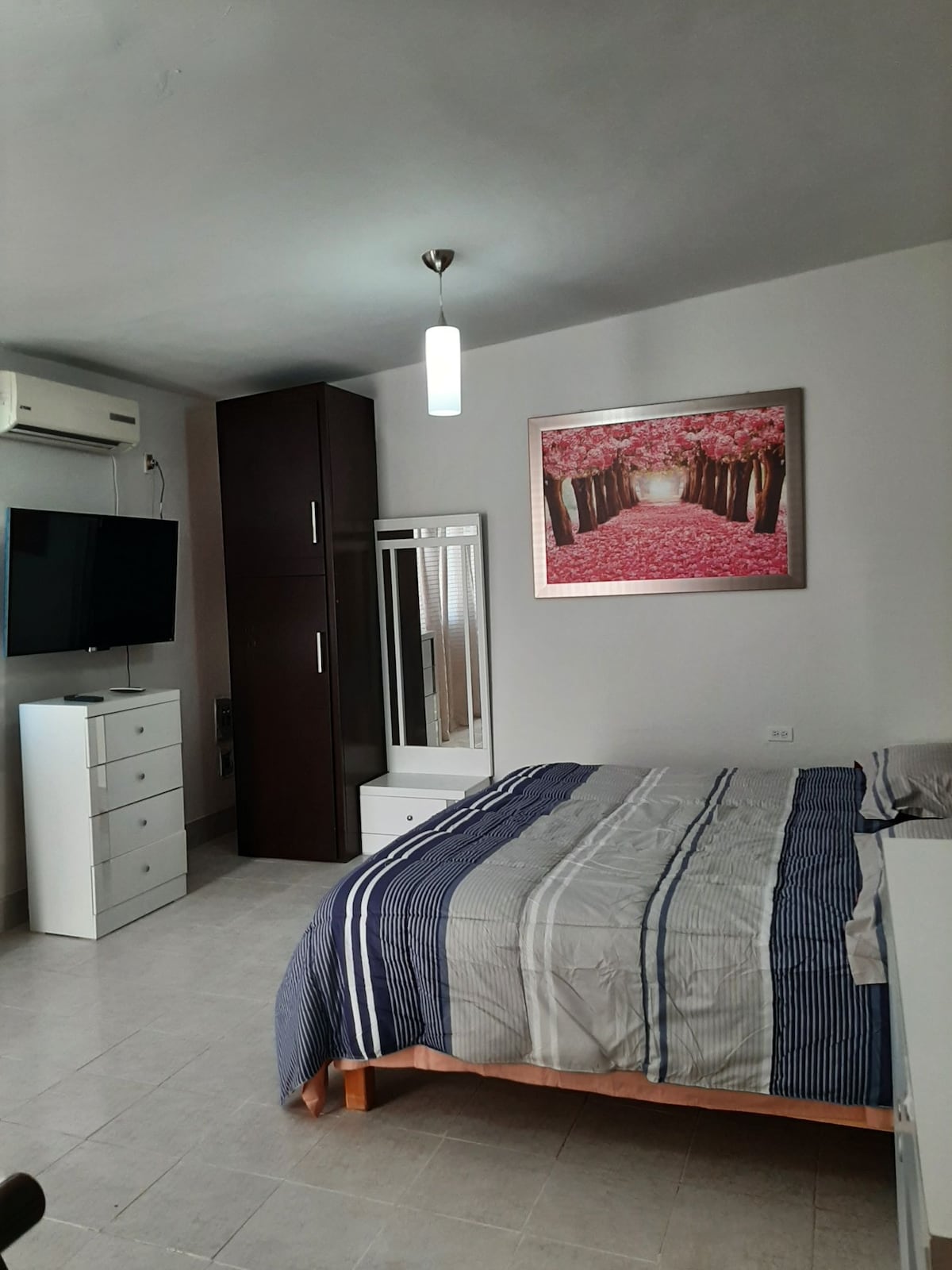 A spacious bedroom features a king-sized bed with a striped blanket, complemented by a decorative wall art depicting a tree-lined path. A television is mounted on the wall, and a tall wardrobe is positioned next to a dresser, providing ample storage space.