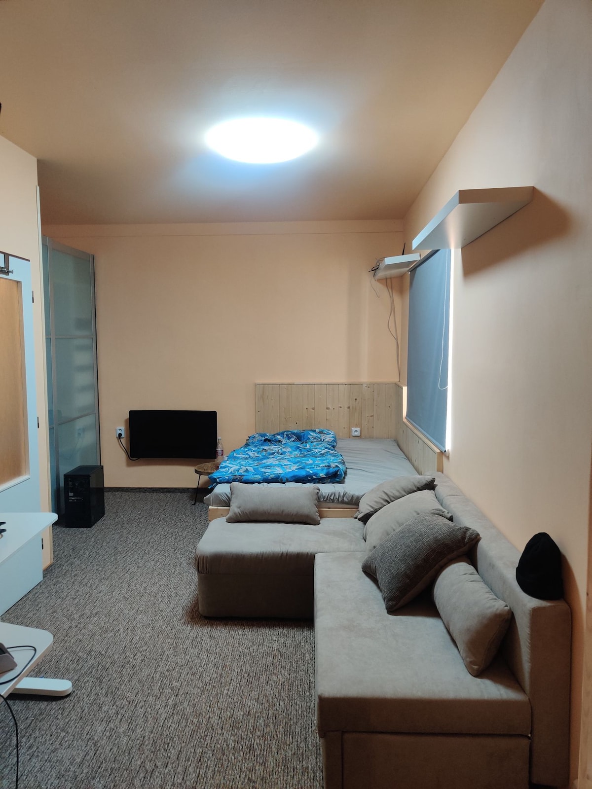 A compact living space features a light-colored couch with plush pillows and a bed dressed in blue bedding. A flat-screen TV is mounted on the wall opposite the bed. Warm ambient lighting is provided by a ceiling fixture, enhancing the room's cozy feel.
