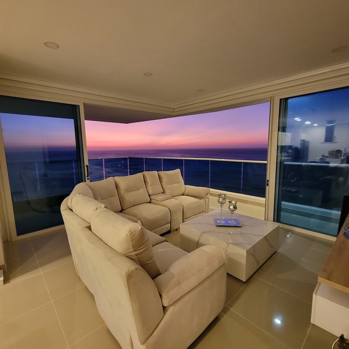 A modern living area features a comfortable light-colored sectional sofa facing expansive glass doors. The outdoor balcony showcases a stunning sunset over the ocean, while a sleek coffee table sits in the center, complemented by elegant glassware.
