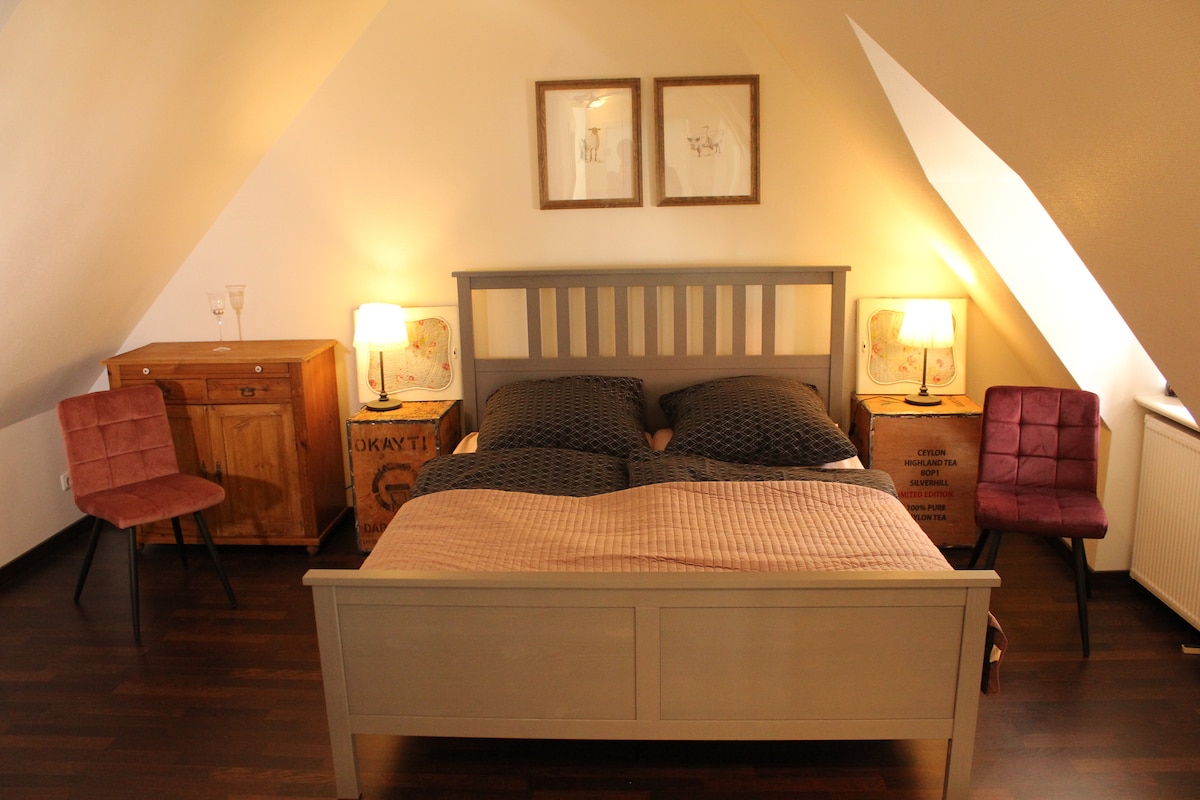 The bedroom features a spacious bed framed in light wood, complemented by soft bedding. Two bedside tables with lamps provide ambient lighting. A wooden dresser is present, and two pink chairs add a touch of color. Angled ceilings create a cozy atmosphere.