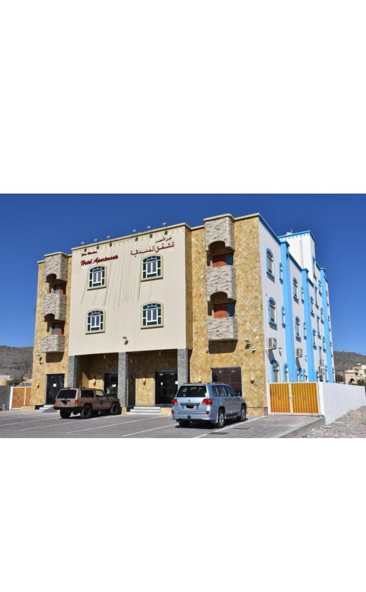 A three-story building features a blend of natural stone and painted exteriors. Large windows are visible on each floor, with balconies providing outdoor space. A parking area is located in front, accommodating multiple vehicles.