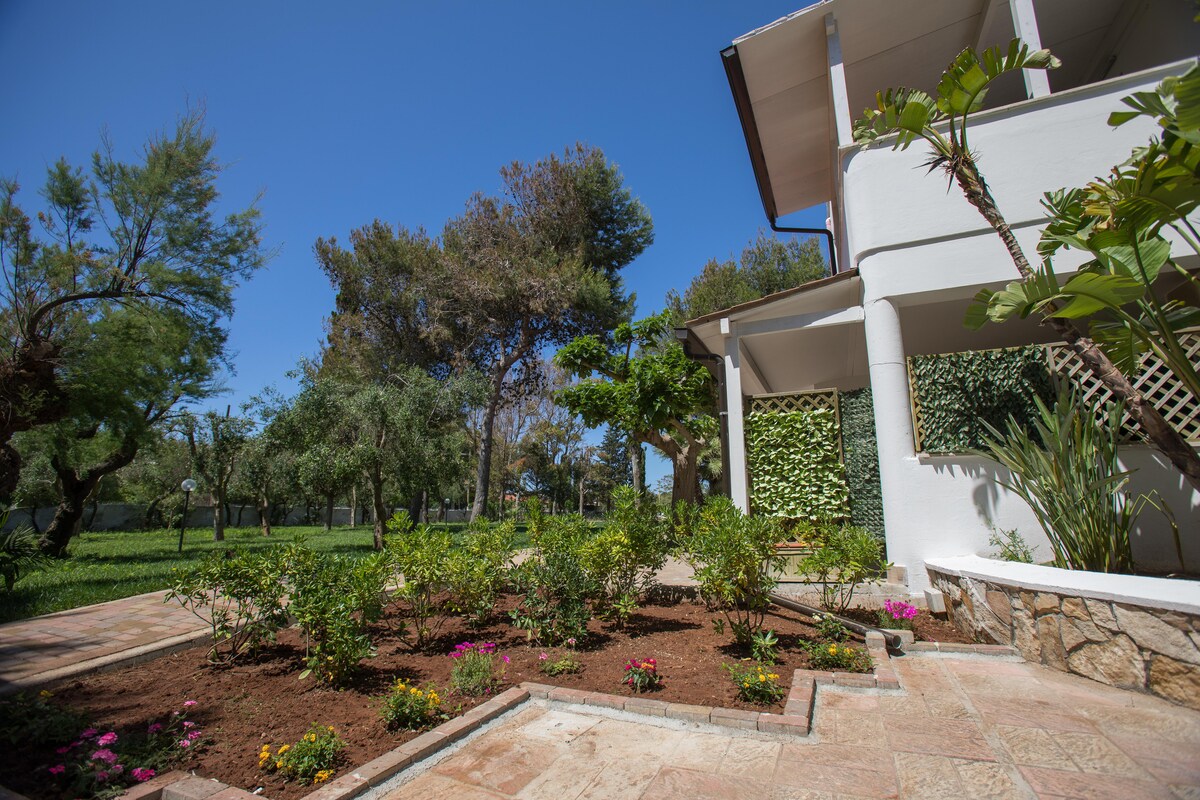 A spacious garden is surrounded by trees, creating a serene outdoor area. Colorful flowers are planted in neat arrangements along the pathways, while a well-kept lawn extends in the background. The structure, featuring large windows, is partially visible among the greenery.