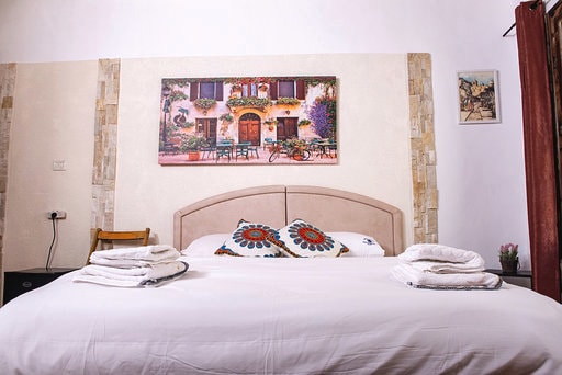 A comfortable sleeping area features a spacious bed adorned with decorative pillows. A large wall art piece depicting a quaint building is displayed above the bed. Soft towels are neatly arranged at the foot of the bed, complementing the overall decor.