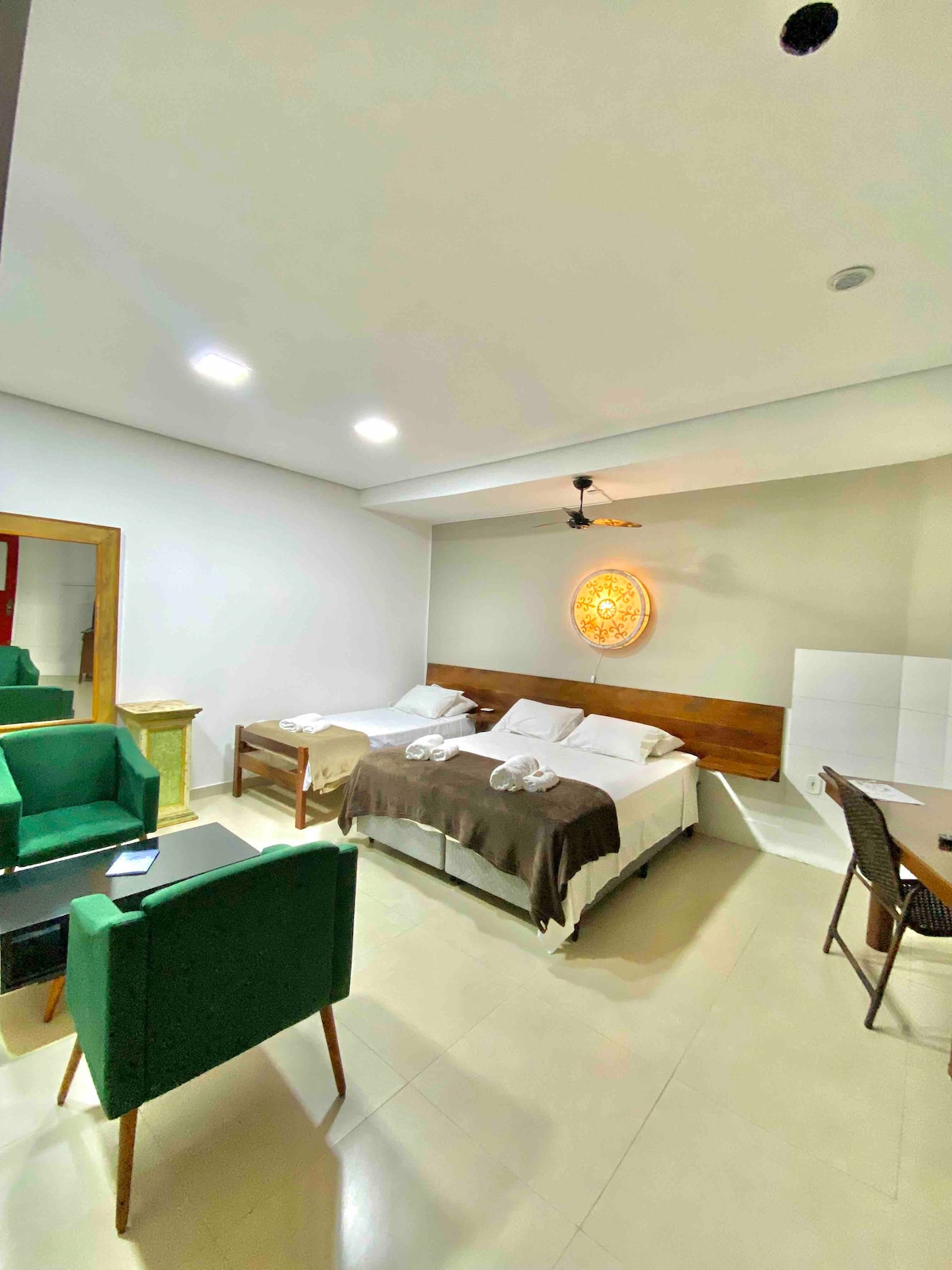 A spacious room is featured with two beds dressed in white linens and dark blankets. A comfortable seating area is provided with green chairs and a small table. Soft lighting illuminates the room, and a decorative wall feature adds subtle warmth.